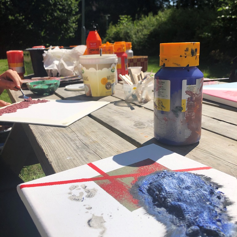 Bilden visar ett uppdukat bord utomhus med färger, penslar och målardukar.