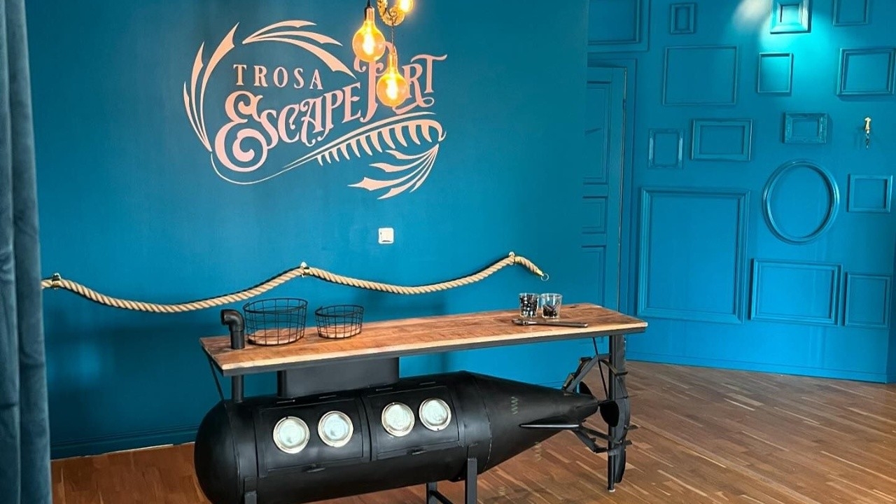 I Escape Port Trosas blåmålade lounge står ett u-båtsliknande bord där fika brukar dukas upp till de gäster som spelar escape roomet. I bakgrunden syns en till blåmålad vägg med tavelramar utan innehåll som blir en del av stukaturen i väggen.