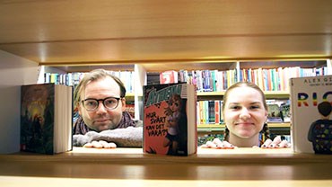 En man och en kvinna kikar leende igenom en bokhylla, där det står tre böcker.