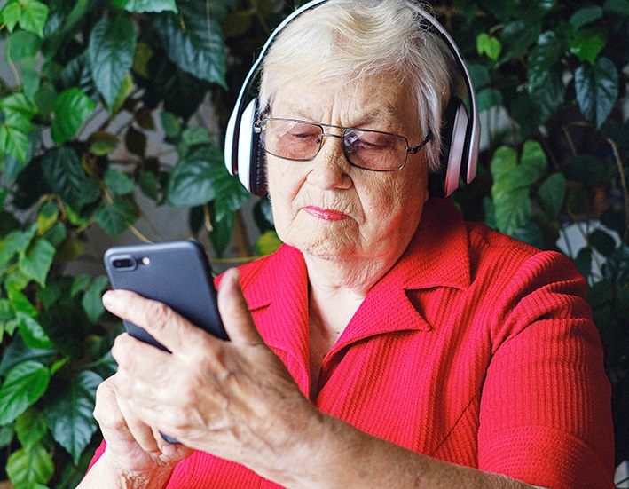 En gammal dam i röd skjorta sitter med en smartphone i handen. Hon har hörlurar. I bakgrunden ser man en grön buske.