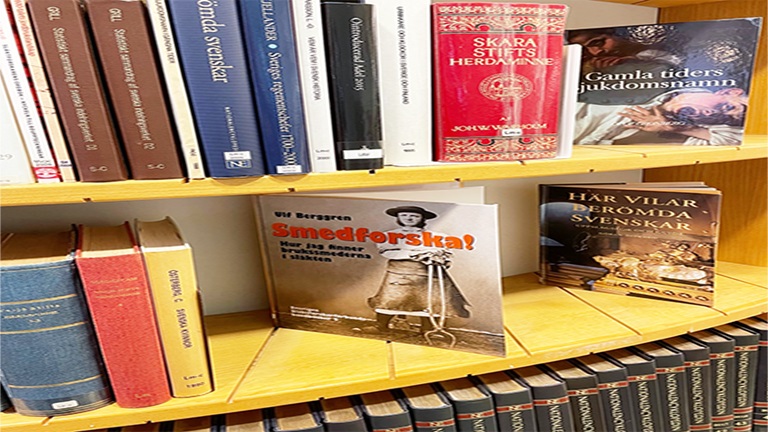 Bokryggar i brunt, vitt, blått, rött och gult i en svängd bokhylla i gulnat trä. Samt tre böcker som handlar om släktforskning som står skyltade med framsidan synlig.