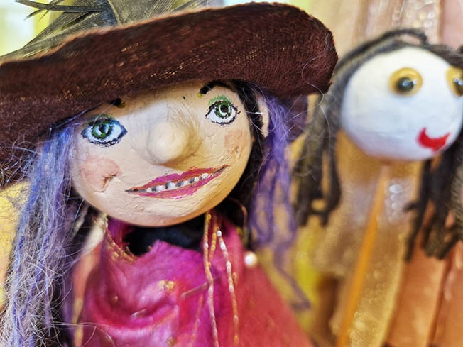 Två dockor mot gulgrå bakgrund. Ena dockan ser ut att bära smink och har lila hår och en brun slokhatt. Den andra dockan är ett vitt huvud med röda läppar och gula ögon och har grått hår.