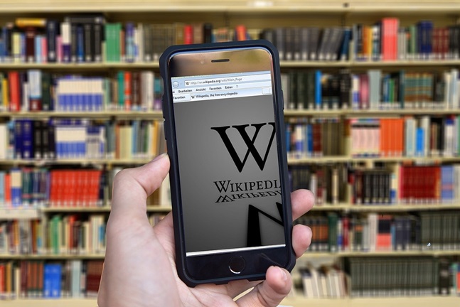 Man ser en hand som håller i en mobiltelefon. På skärmen visas Wikipedias svenska hemsida. I bakgrunden stor flera bokhyllor fyllda med böcker.