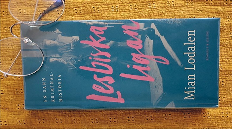 En närbild på boken Lesbiska Ligan av Mian Lodalen, samt ett par ihopfällda, stålbågade glasögon som ligger ovanpå boken i vänstra hörnet.
