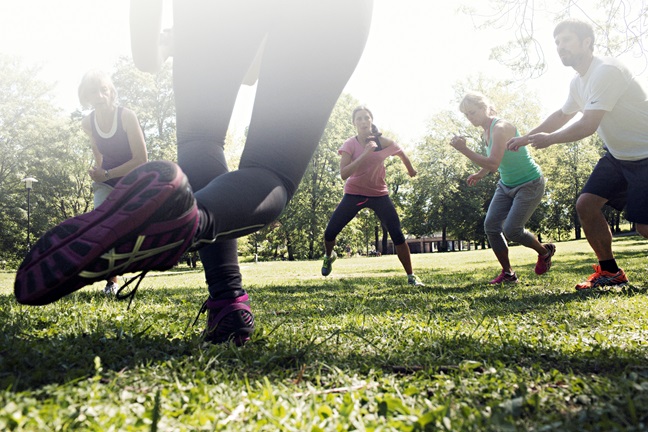 På bilden ses ett gäng glada motionärer som tränar utomhus på ett grönt fält. Aktiviteten är jympa. 