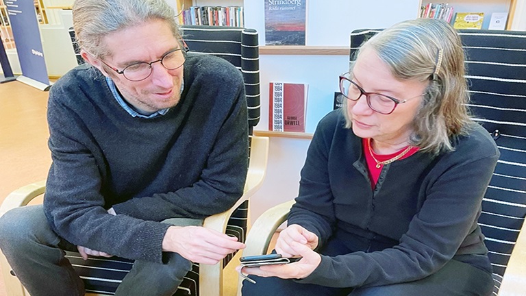 En man och en kvinna sitter bredvid varandra i varsin fåtölj. Båda lutar sig över mobiltelefonen som kvinnan håller i sin hand. De pekar och tittar på telefonen.