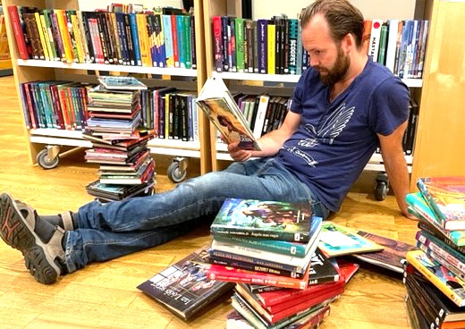 En man sitter på golvet framför en fylld bokhylla djupt försjunken i en bok. Framför mannen ligger högar med böcker.