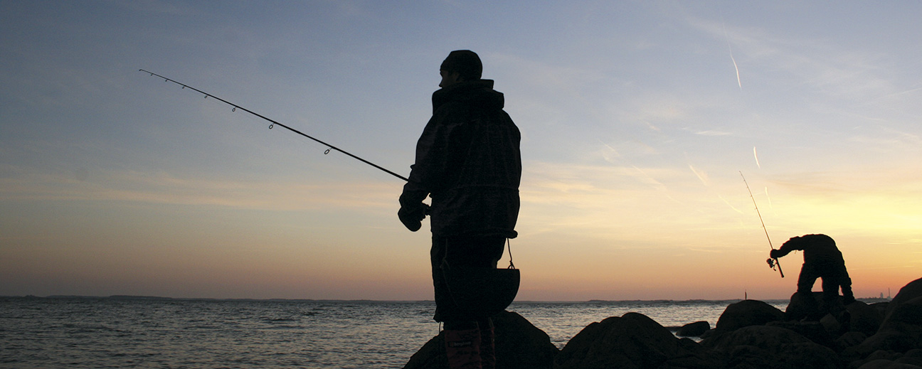 Två fiskare står på klippor i solnedgången.