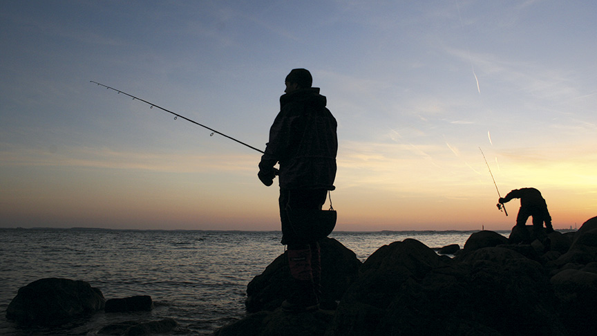 Två fiskare står på klipporna i motljus i solnedgången.
