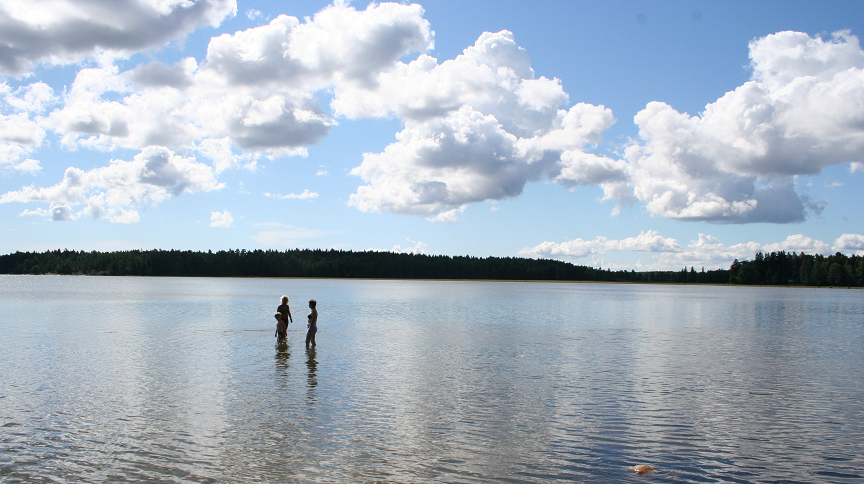Två vuxna och ett barn står ute i vattnet vid en badplats. 